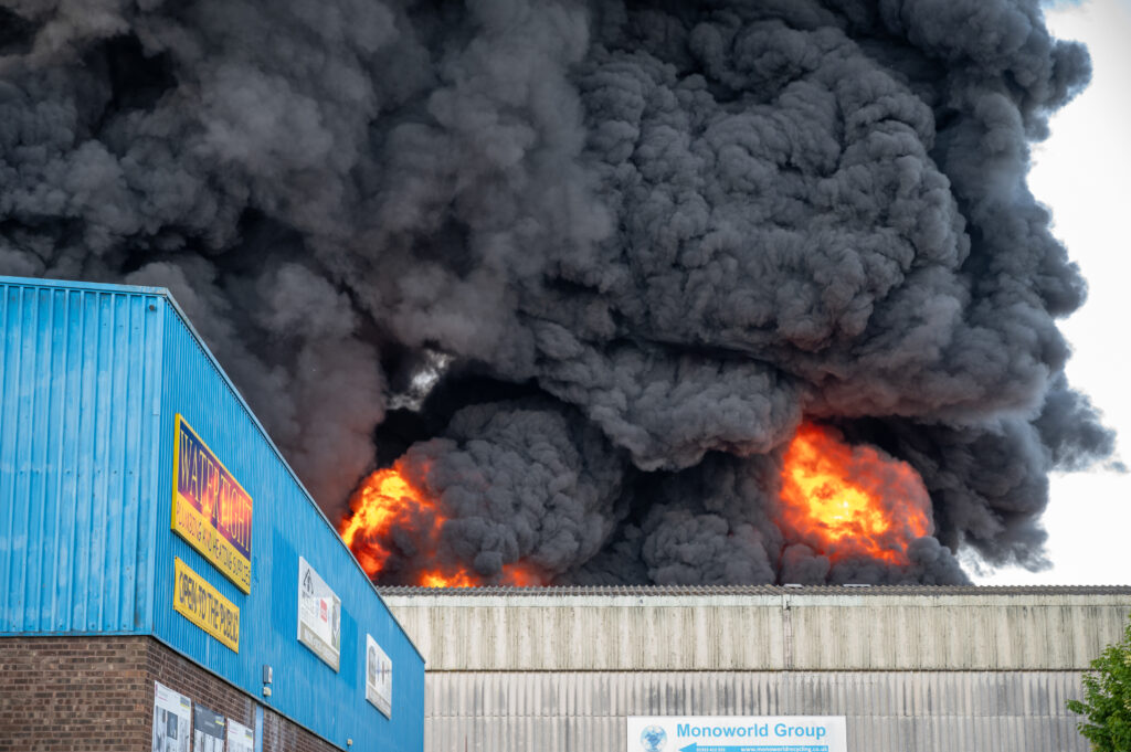 Large industrial fire with thick black smoke and bright flames rising behind a blue-sided building labeled 'Water Tight Plumbing & Drainage Supplies' and 'Monoworld Group