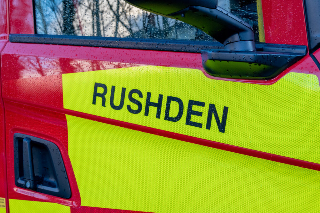 Side view of a red and yellow emergency vehicle labeled 'RUSHDEN', part of the fire and rescue service