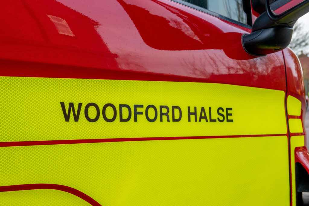 Side view of a reflective emergency vehicle with 'WOODFORD HALSE' written in black capital letters on a yellow and red background, partially showing a side mirror.