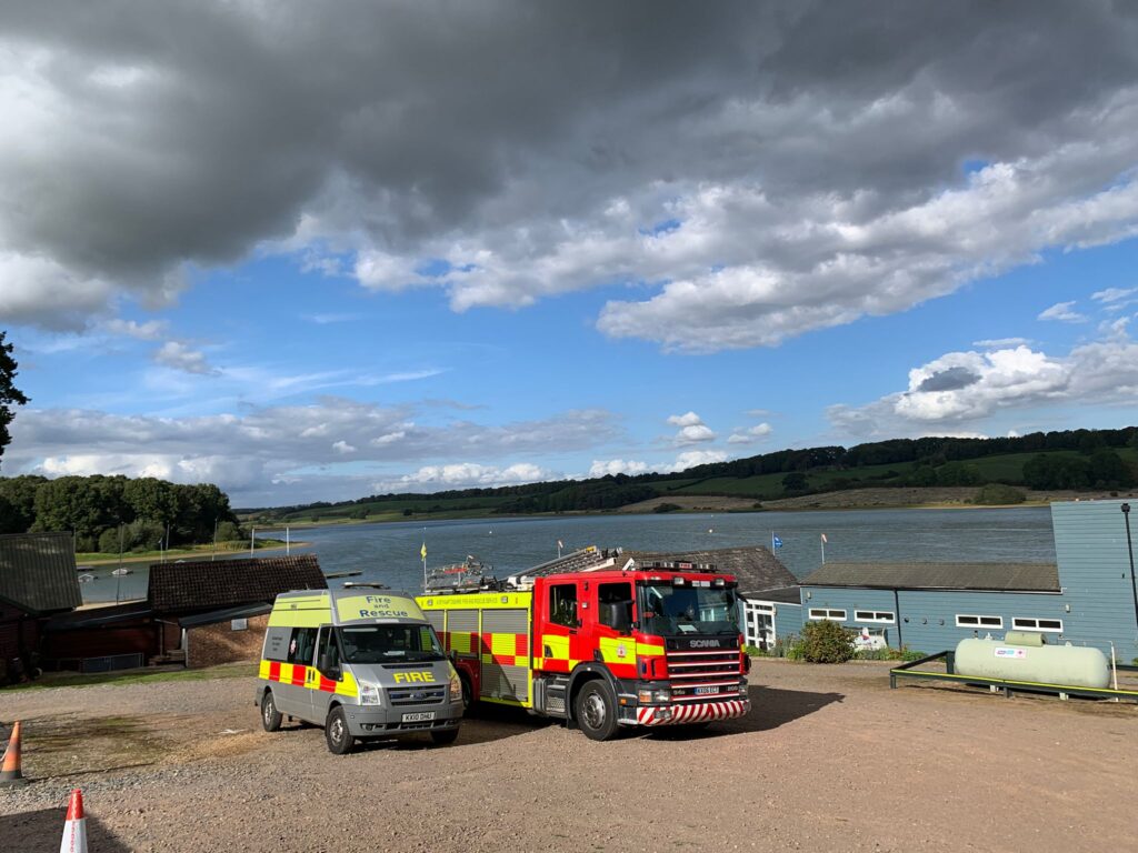 Two emergency vehicles—a yellow fire van marked "FIRE" and a red fire truck—are parked near a large body of water under a cloudy sky. Buildings line the water’s edge with rolling hills in the background.
