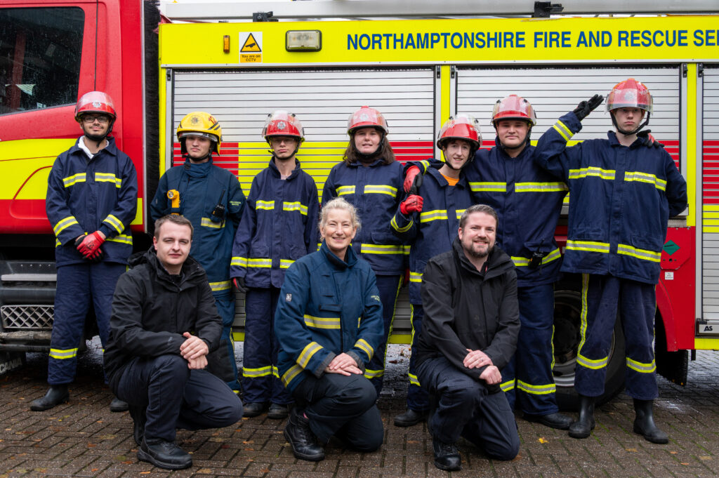 A group of individuals wearing dark blue fire service uniforms with reflective yellow stripes and red helmets are posing in front of a fire engine. The fire engine has a bright yellow and red design with visible text reading 'Northamptonshire Fire and Rescue Service'. Three people are kneeling in the front row while the others stand behind them. The ground is paved, and the background shows part of the fire engine’s side compartments.