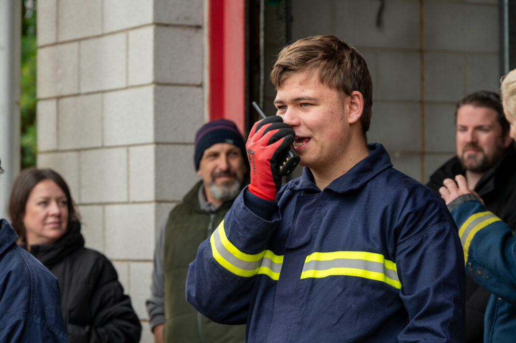 A person wearing a dark blue fire service uniform with reflective yellow stripes and red gloves is holding a handheld radio to their mouth, appearing to communicate. The individual is standing in front of a partially open garage or building entrance with a concrete block wall. Several other people in similar attire and casual clothing are visible in the background, as part of a training scenario.