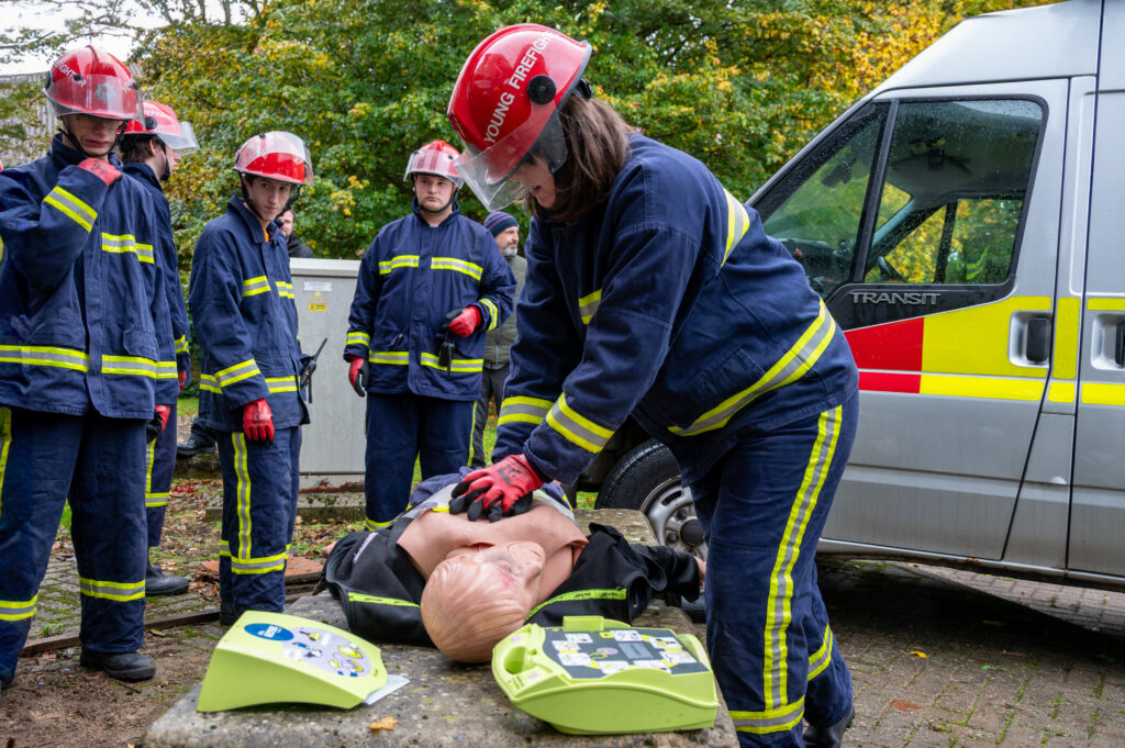 A group of individuals wearing dark blue fire service uniforms with reflective yellow stripes and red helmets are gathered outdoors near a silver emergency vehicle with red and yellow markings. One person is performing chest compressions on a CPR training mannequin placed on a stone surface, with a green automated external defibrillator (AED) unit positioned nearby. The scene appears to be part of a first aid or emergency response training exercise. Trees with autumn foliage are visible in the background.