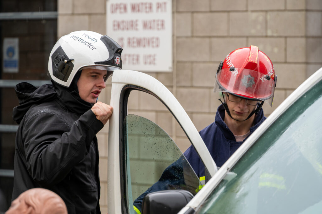 Two individuals wearing fire service helmets are standing next to a white vehicle. One helmet is white with the words 'Youth Instructor' printed on it, and the other is red. The person with the white helmet is leaning on the open door of the vehicle, while the other stands nearby. In the background, there is a light gray brick wall with a sign that reads “OPEN WATER PIT ENSURE WATER REMOVED AFTER USE.”