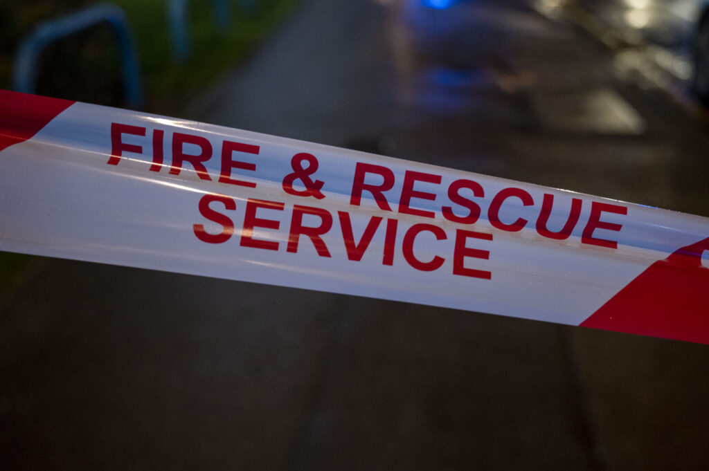 Close-up of a red and white barrier tape with the words “FIRE & RESCUE SERVICE” printed in bold red letters. The tape is stretched across a dimly lit outdoor scene, with a blurred pavement and faint blue lights visible in the background.