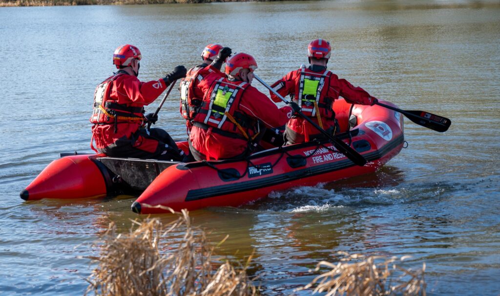 A group of four individuals in red protective gear and helmets paddle a red inflatable rescue boat labeled "FIRE AND RESCUE" across a body of water. They are equipped with life vests and paddles.