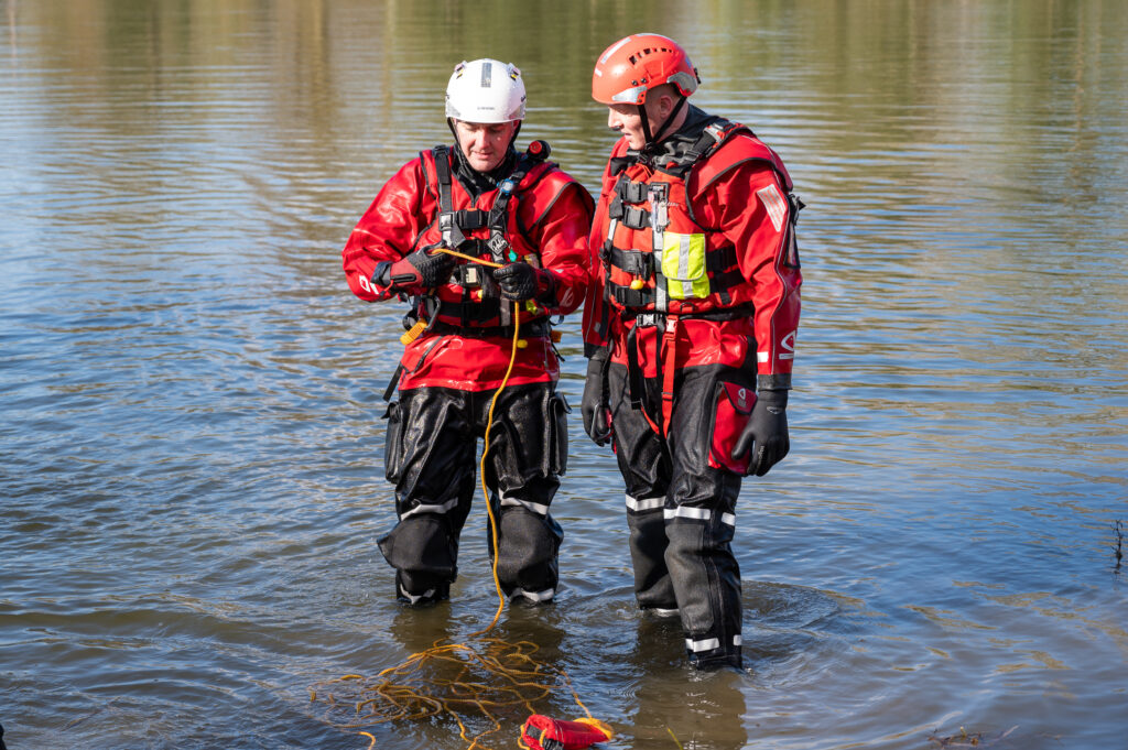 Two individuals in red protective gear and helmets stand knee-deep in water, managing a yellow rope. Both wear life vests, gloves, and waterproof suits.