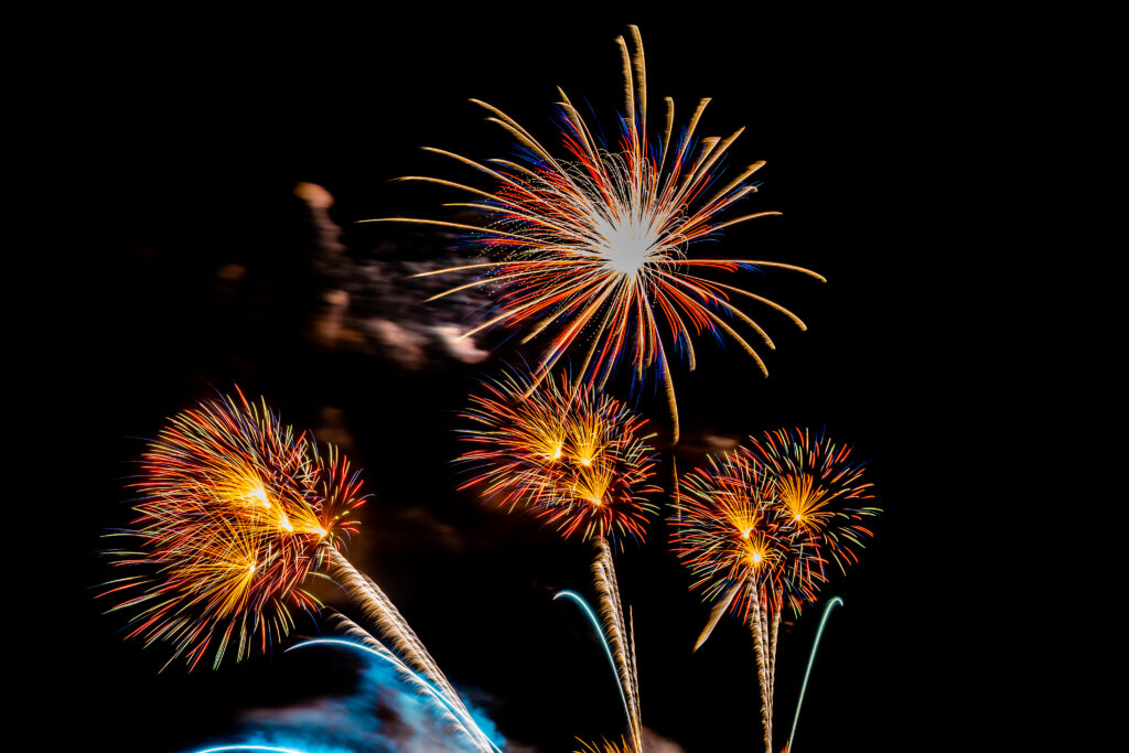 Bright, colorful fireworks exploding against a dark night sky. The display features multiple bursts in shades of red, orange, yellow, and blue, with streaks of light radiating outward and faint smoke visible in the background.