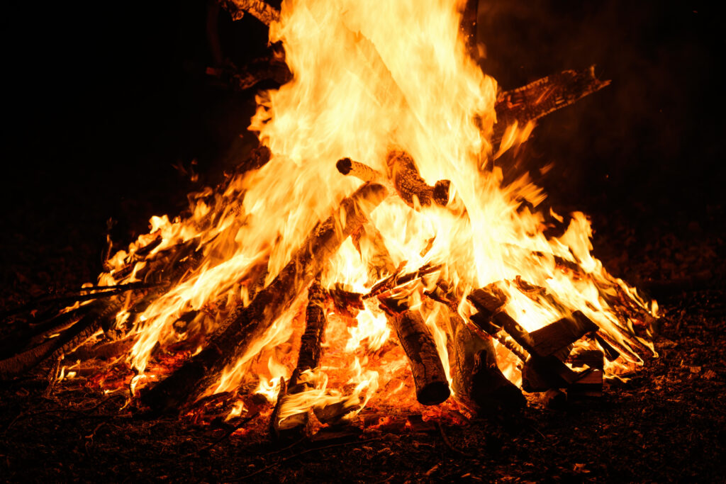 Large bonfire burning brightly at night, with thick logs stacked in a pile and intense orange flames rising high against a dark background.