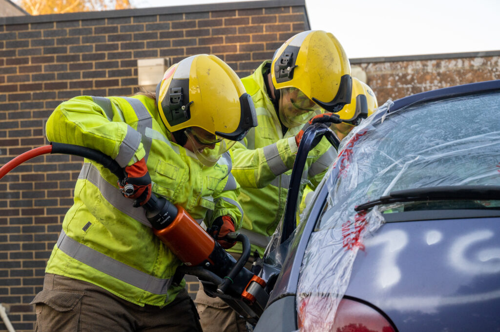Two firefighters wearing yellow helmets and high-visibility protective gear use hydraulic cutting equipment to remove the door of a damaged car during a rescue training exercise. The car is partially covered with clear protective plastic, and the background shows a brick wall.
