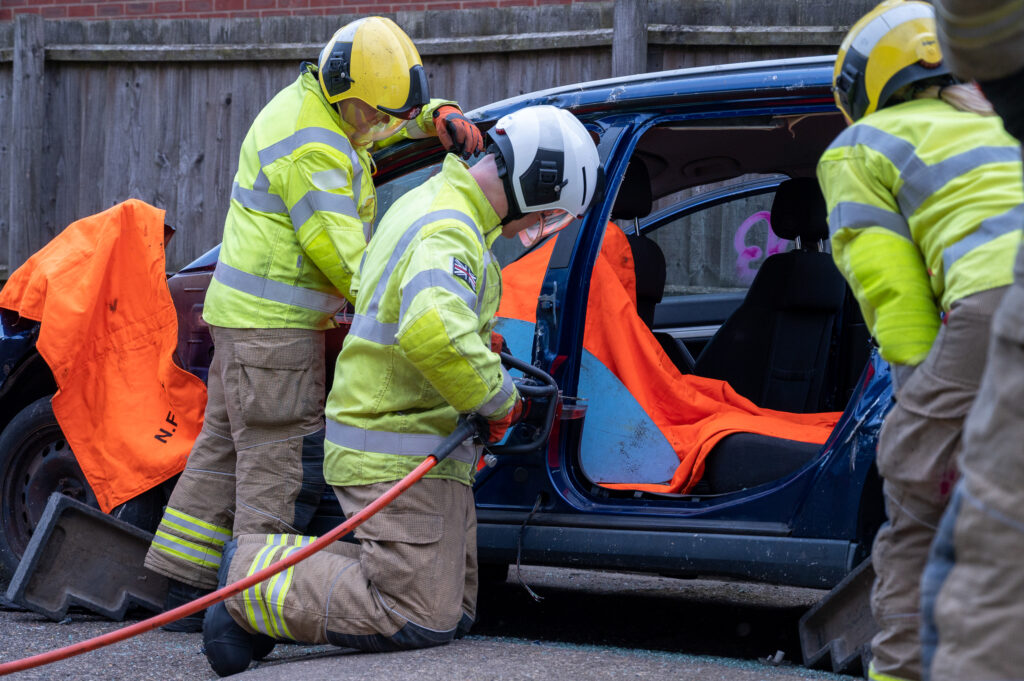 Three firefighters in high-visibility protective gear work on a dark blue car during a rescue training exercise. The car doors are open, and bright orange safety sheets cover the interior. One firefighter kneels using hydraulic cutting equipment on the car frame, while others assist nearby. The scene is outdoors, with a wooden fence in the background.