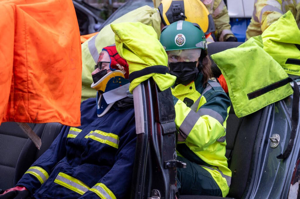 Emergency responders assist a person seated inside a damaged vehicle during a rescue operation. The individual is wearing a neck brace and protective clothing, while bright safety sheets and rescue equipment surround the car interior.