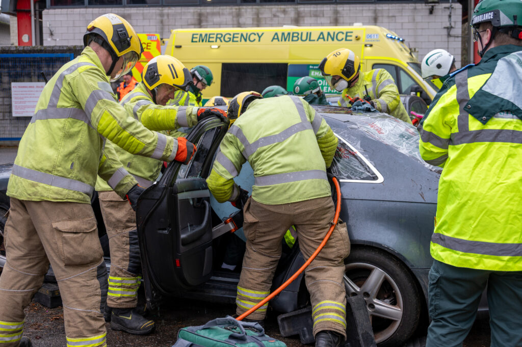 Firefighters in high-visibility protective gear use hydraulic rescue tools to cut open the door of a damaged car during an emergency response exercise. Several emergency personnel are working around the vehicle, and an ambulance is parked in the background.