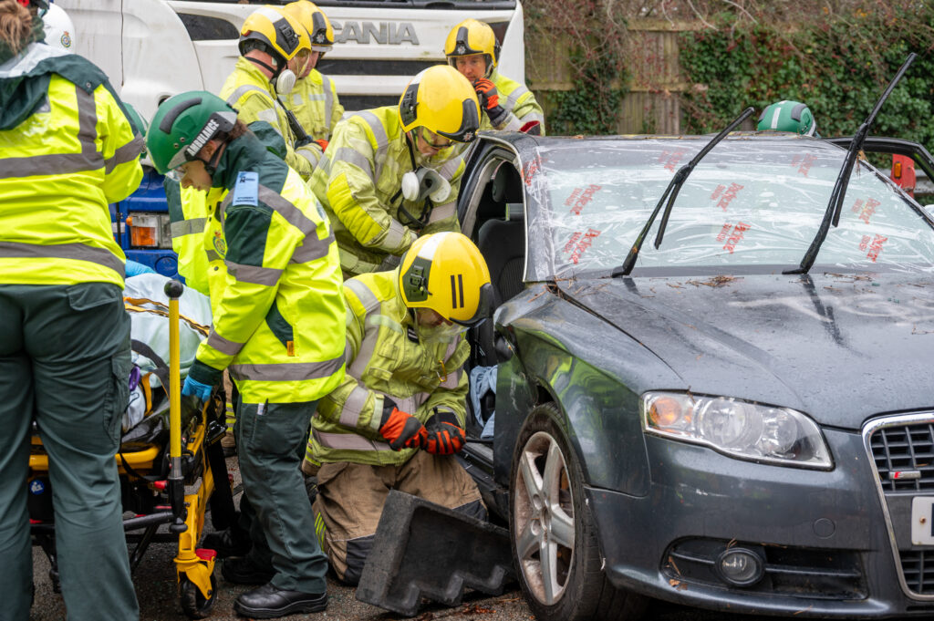 Emergency responders work together to rescue an occupant from a damaged car. Firefighters in yellow helmets and protective gear kneel by the open door, while paramedics in green and yellow jackets prepare a stretcher nearby. The car’s windshield is shattered and partially removed, and rescue equipment is visible on the ground