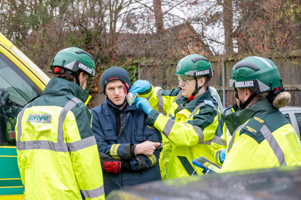 Three paramedics in green and yellow high-visibility jackets and helmets assist a person wearing a dark protective suit near an ambulance. One paramedic is adjusting a medical collar on the individual while others stand close by, providing support.