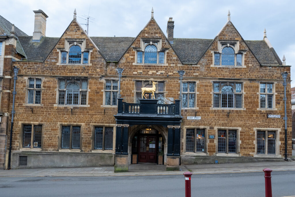 Historic stone building with ornate architecture, featuring multiple pointed gables, large mullioned windows, and a central entrance with a black balcony topped by a golden hind statue. The building has signs reading ‘The Hind Hotel’ and is situated along a paved street with two red bollards in the foreground.