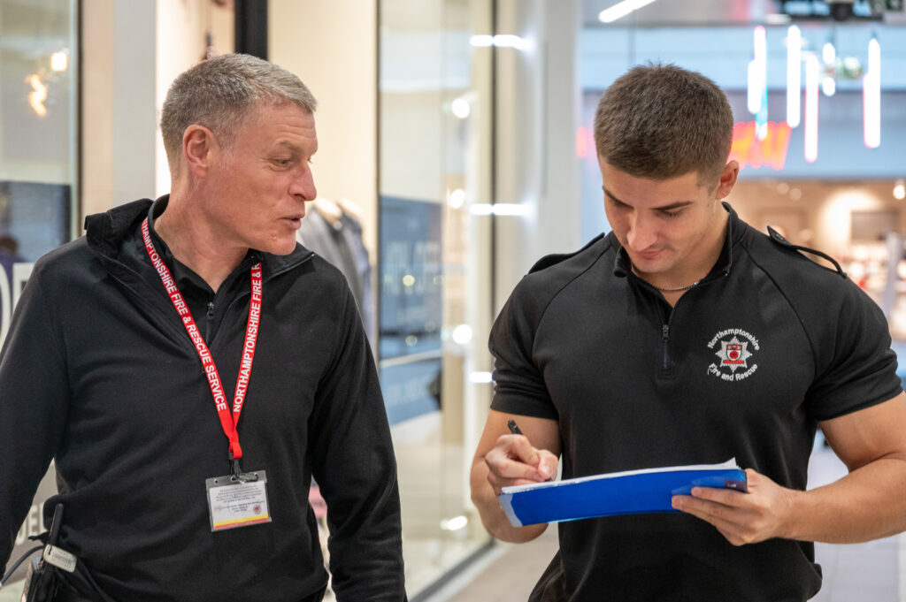 Two firefighters are inside a shopping centre. One is holding a blue clipboard and writing notes, while the other stands beside, wearing a lanyard and ID badge. Bright store fronts and glass panels are visible in the background.