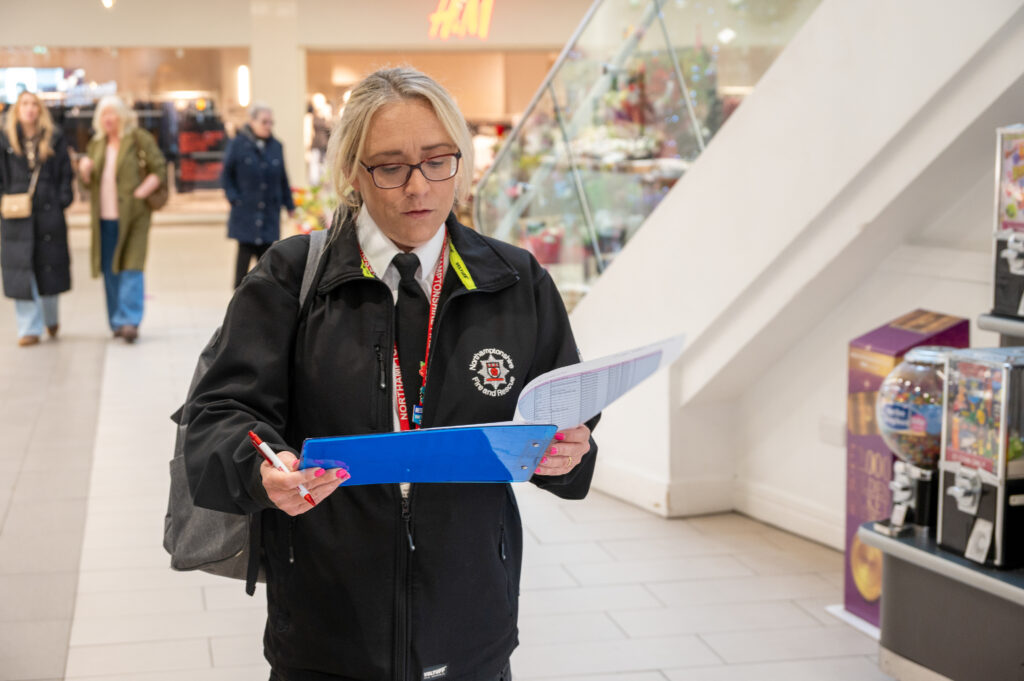 A fire and rescue service Protection Officer wearing a black uniform and lanyard is standing inside a shopping centre, holding a blue clipboard and a pen while reviewing a checklist. There are shoppers and retail displays in the background.