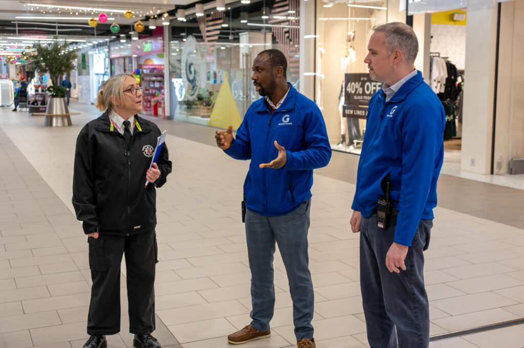 A fire and rescue service Protection Officer is speaking with two staff members from the Grosvenor Centre inside a shopping centre. The fire officer is holding a clipboard, while the shopping centre staff wear blue jackets and have radios clipped to their belts. Shops and festive decorations can be seen in the background.