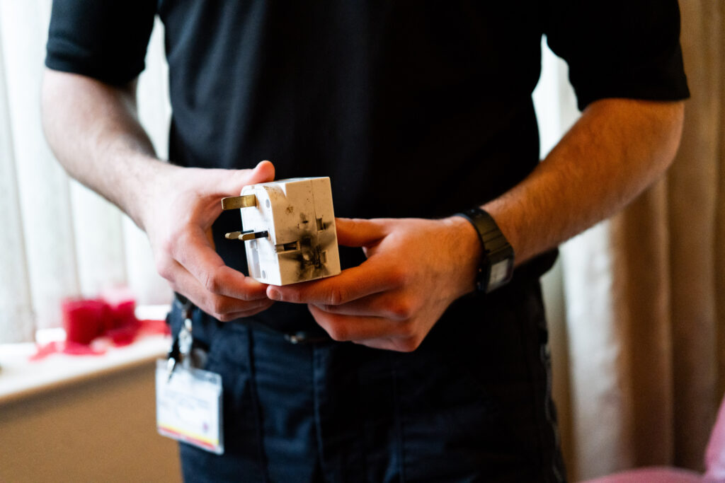 A person wearing a dark shirt holds a damaged electrical plug with burn marks on its surface. The individual&rsquo;s ID badge hangs from their waistband, and the scene appears to take place indoors near a window with soft light coming through.