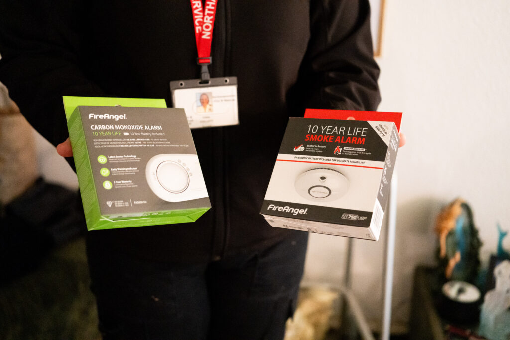 A person wearing a black uniform and lanyard holds two boxed FireAngel safety devices: a carbon monoxide alarm in green packaging and a 10‑year life smoke alarm in red and white packaging