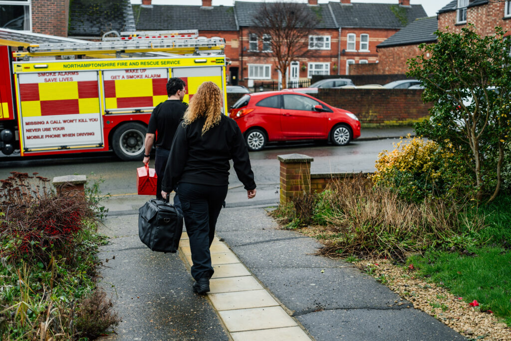 Two uniformed individuals walk down a garden path carrying equipment bags toward a parked fire engine on a residential street. A red car is parked nearby, and houses line the background on a damp, overcast day.