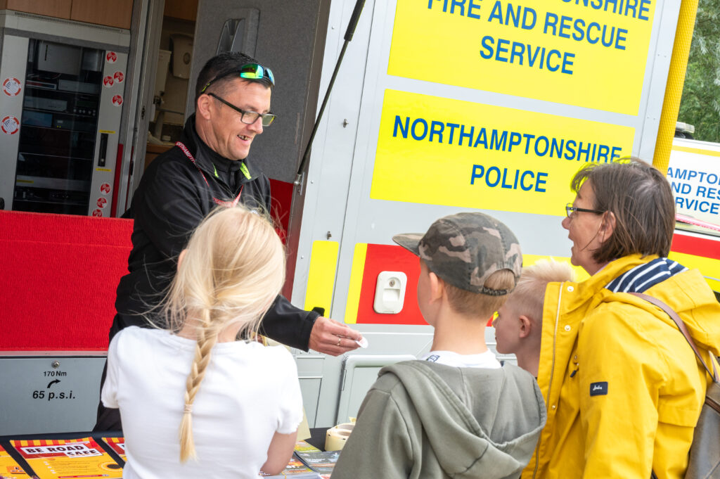 A person wearing a black uniform and sunglasses is speaking to a group of children at an outdoor event. The children are standing in front of a display table with safety information and leaflets. Behind the person is a large emergency service vehicle with bright yellow signage that reads &ldquo;FIRE AND RESCUE SERVICE&rdquo; and &ldquo;NORTHAMPTONSHIRE POLICE.&rdquo; The scene suggests a community safety or public engagement event.