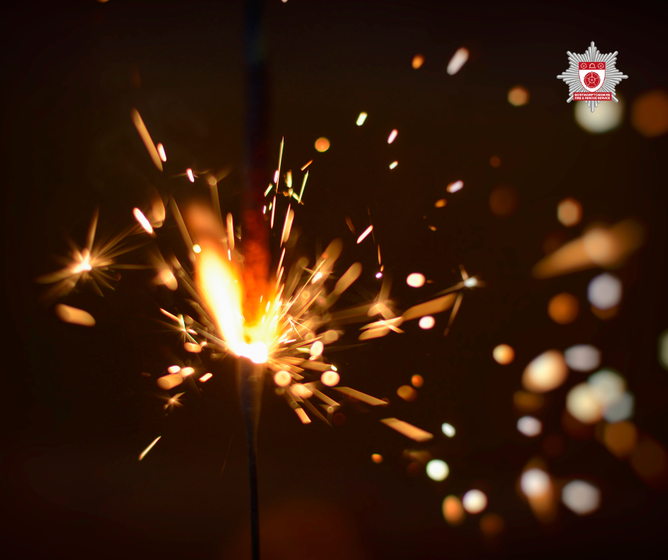 Close-up of a lit sparkler emitting bright orange and yellow sparks against a dark background, with a fire and rescue service badge logo in the top right corner.