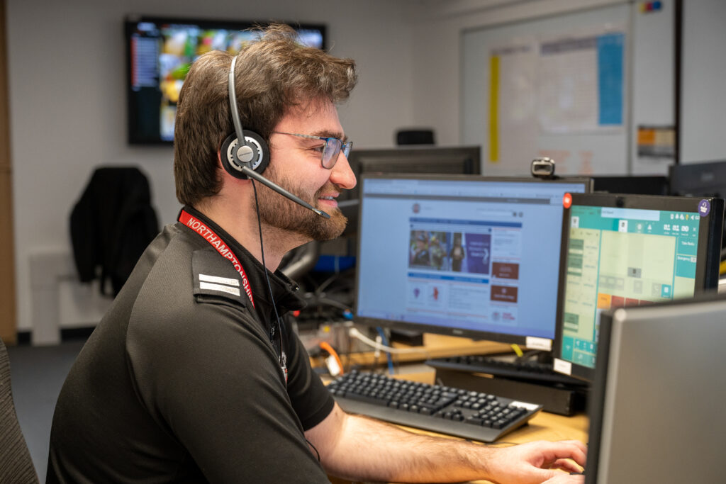 A control room operator wearing a headset works at a desk with multiple monitors. The screens show a mix of a website, mapping tools, and communication software. A keyboard, mouse, and other office equipment are arranged on the desk, with additional monitors and screens visible in the background.