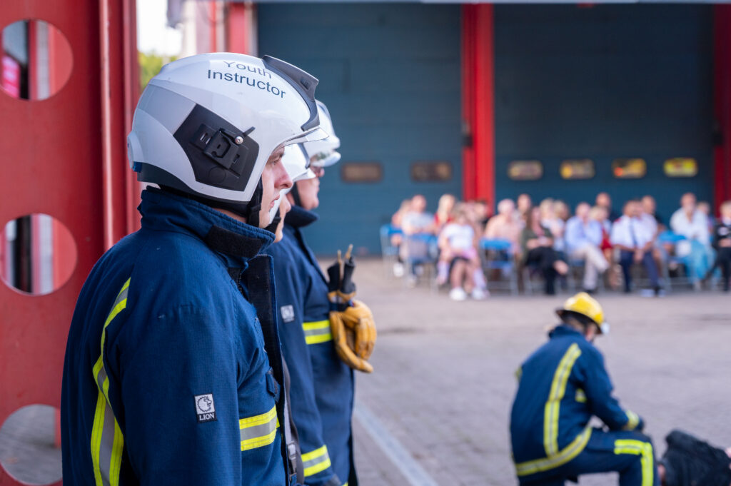 Several individuals in firefighting protective clothing stand and kneel in front of a fire station. One person in the foreground wears a helmet labeled &ldquo;Youth Instructor&rdquo; and looks toward a seated audience in the background. The group of spectators sits outside the station&rsquo;s open bays, watching the activity taking place.