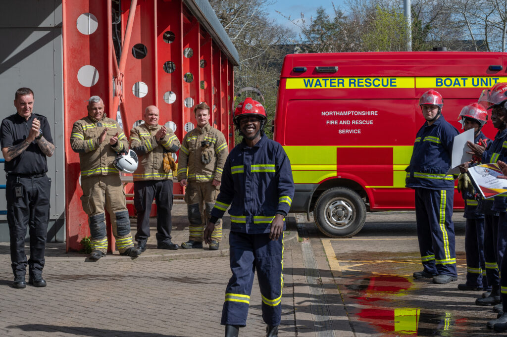 A firefighter in protective clothing and a red helmet walks forward during a training exercise while colleagues stand applauding on either side; a red water rescue vehicle marked ‘Northamptonshire Fire and Rescue Service’ is parked nearby.