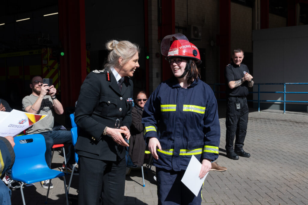 Firefighters in protective clothing and helmets carry a simulated casualty during a training exercise outside a fire station building, with other personnel and equipment visible nearby.