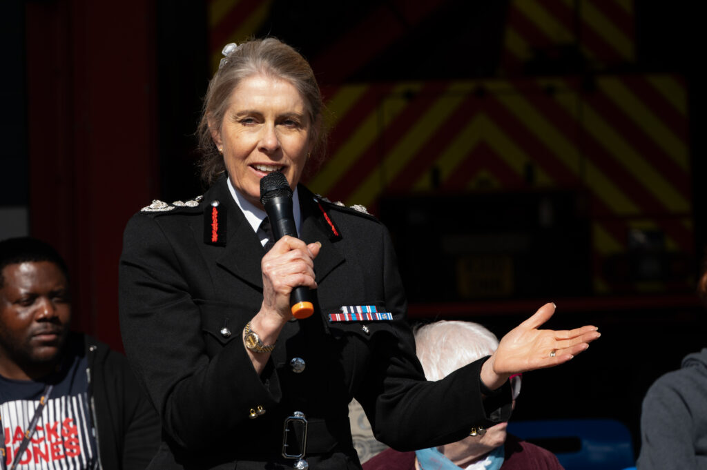 A uniformed fire service representative speaks into a handheld microphone during an outdoor event, standing in front of fire station bays while people sit listening in the background.