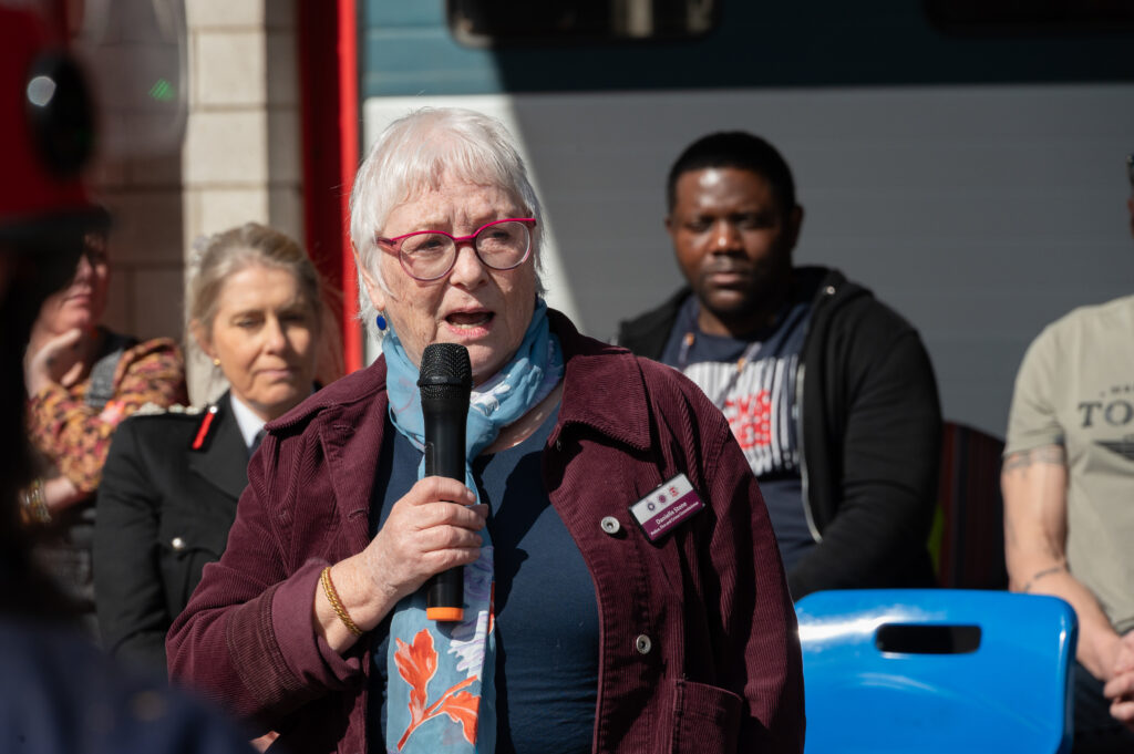 An older person with short grey hair and glasses speaks into a handheld microphone while seated listeners and fire service personnel sit behind them outdoors, in front of a fire station.