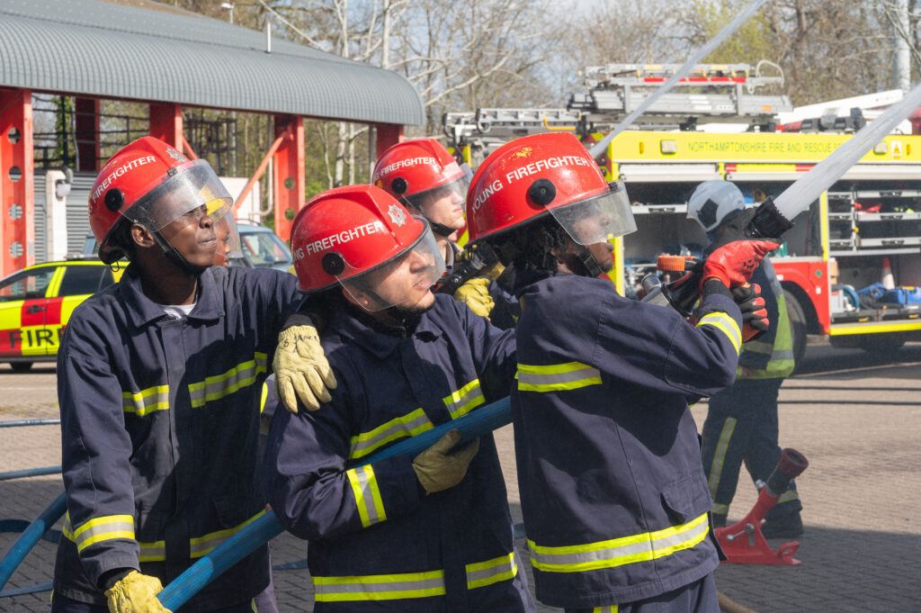 Firefighters wearing protective gear and red helmets practise handling a high‑pressure hose during a training exercise, with a fire engine and training building visible in the background.