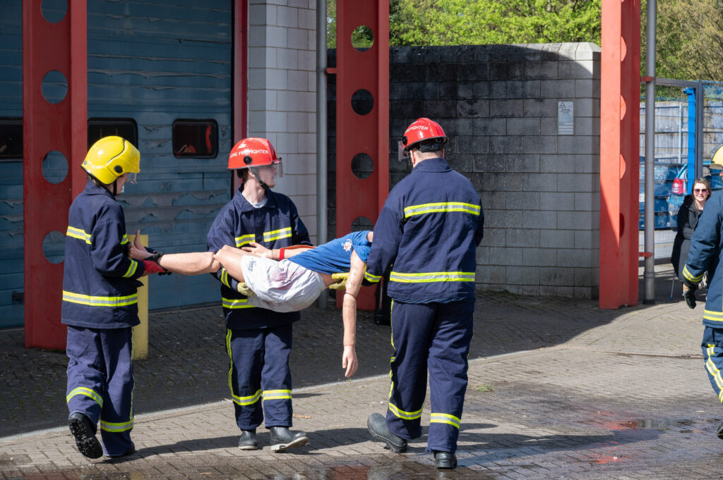 Firefighters in protective clothing and helmets carry a simulated casualty during a training exercise outside a fire station building, with other personnel and equipment visible nearby.