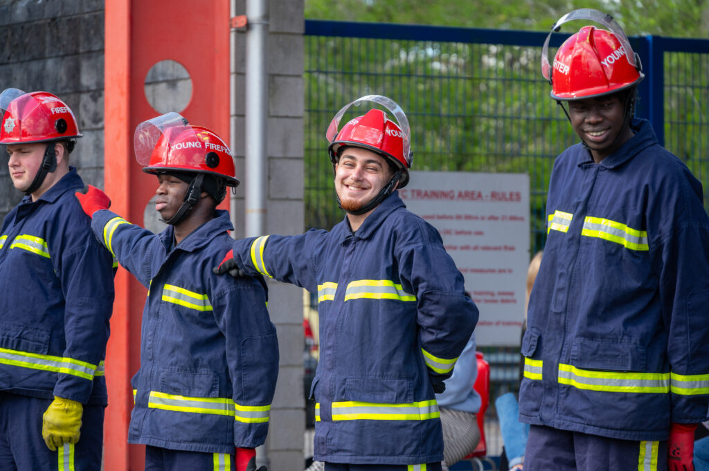 A group of firefighters in protective clothing and red helmets stand together outdoors during a training session, with one trainee facing the camera while others look to the side near a training area fence