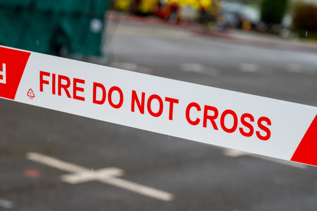 A small fire burning on the ground at night, with bright yellow flames rising from a bundle of dry straw and twigs on patchy grass, surrounded by embers and faint smoke against a dark outdoor background.