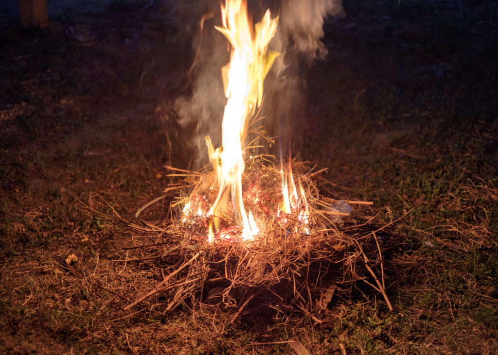 A small fire burning on the ground at night, with bright yellow flames rising from a bundle of dry straw and twigs on patchy grass, surrounded by embers and faint smoke against a dark outdoor background.