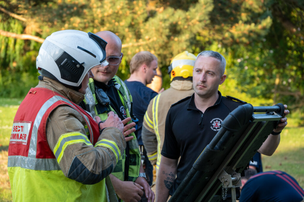 Firefighters in protective clothing and helmets discuss a plan outdoors, standing beside equipment while other crew members and trees are visible in the background.