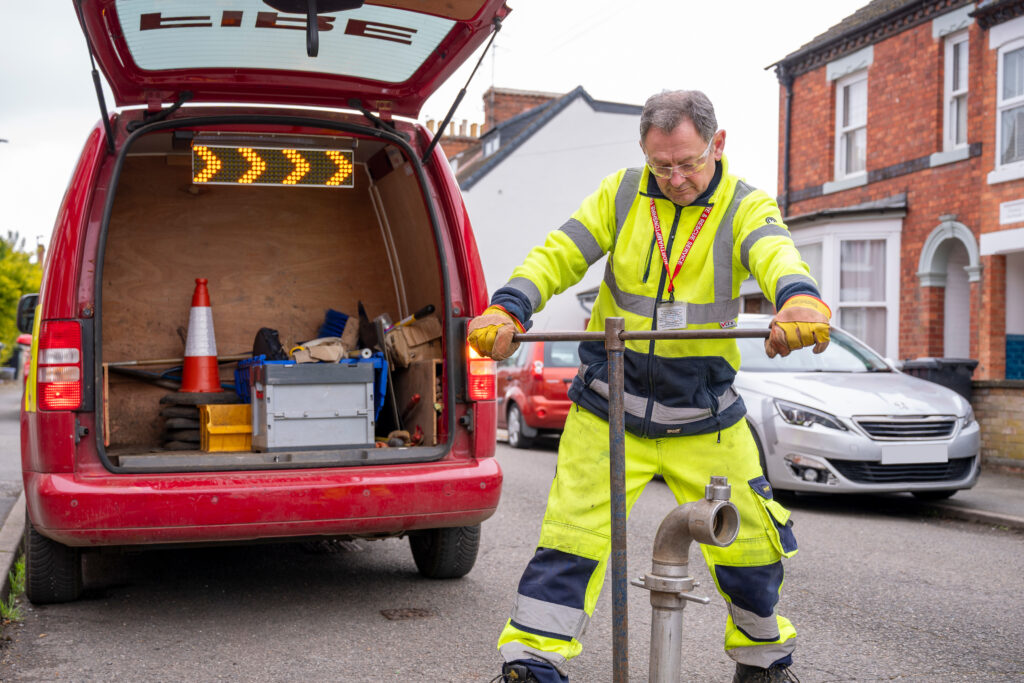 High‑visibility utility worker operating a street fire hydrant with a wrench beside a red service van parked on a residential road