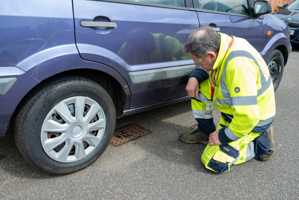 High‑visibility utility worker kneeling beside a parked car, inspecting a small fire hydrant set into the tarmac next to the vehicle&rsquo;s front wheel