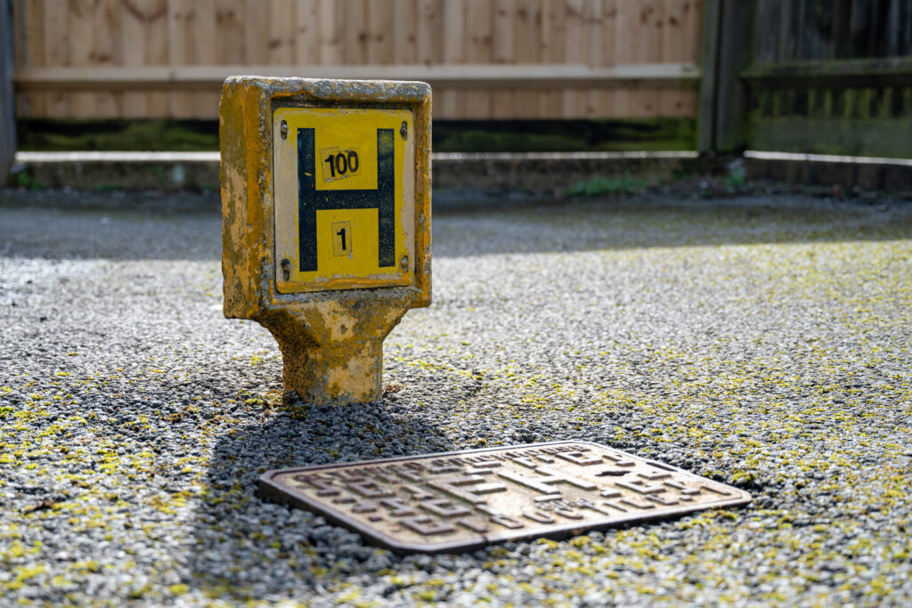 Close-up of a yellow fire hydrant marker post labelled &lsquo;H 100 / 1&rsquo; next to a metal street drain on a paved surface.