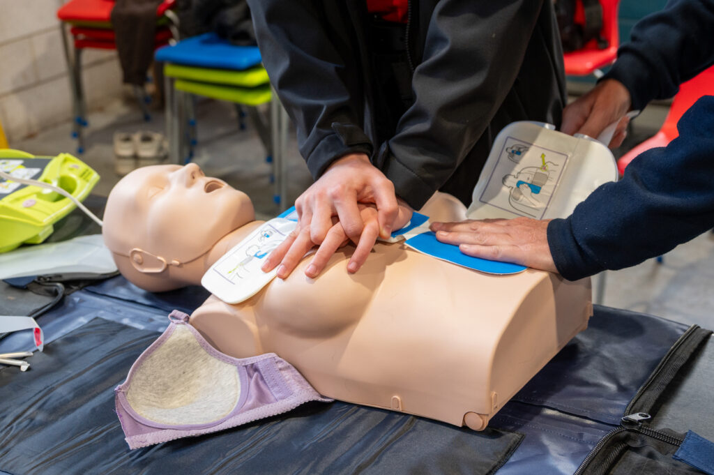 Close-up of hands performing chest compressions on a CPR training manikin while another person positions defibrillator pads on the manikin’s chest.