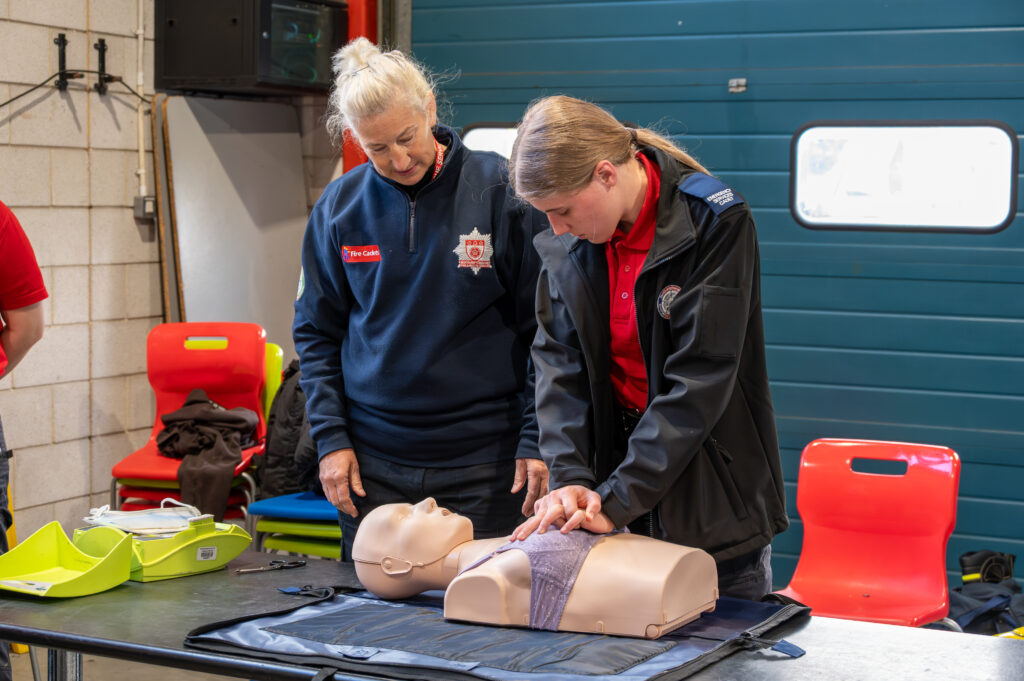 Two fire service trainees practice CPR on a training manikin while an instructor observes and guides, in a fire station training room with chairs and equipment in the background.