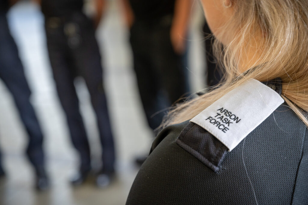 Close-up of the back of a uniformed person with a shoulder tab reading &ldquo;Arson Task Force,&rdquo; with several blurred figures standing in the background.