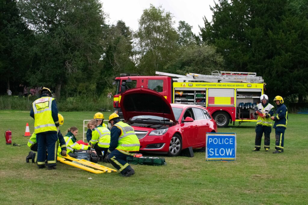 Firefighters conducting a rescue demonstration in a grassy park, treating a casualty beside a red car with its bonnet open, while a Northamptonshire Fire and Rescue Service engine and a “POLICE SLOW” sign are visible nearby.
