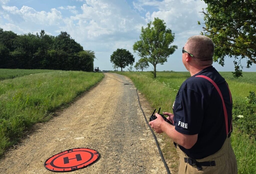 A firefighter stands on a rural dirt track beside green fields and trees, holding a remote controller and looking toward the sky. A circular red landing pad with a large “H” lies on the ground nearby, with a long hose stretched along the path under a bright, partly cloudy sky.