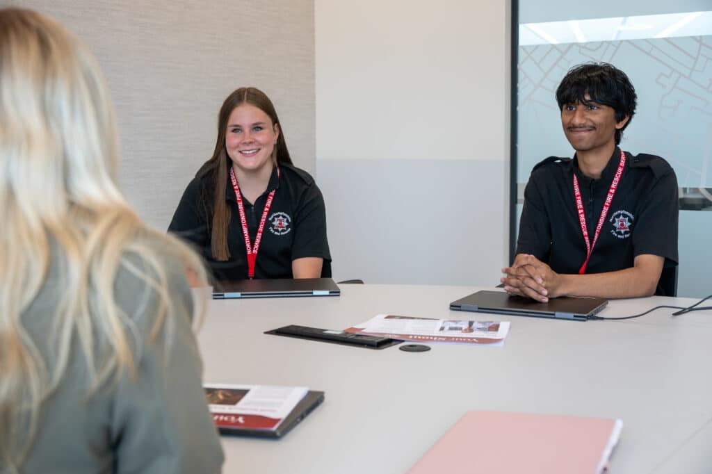 Two Northamptonshire Fire and Rescue Service interns sit at a meeting table with laptops, taking part in a discussion with another person in a meeting room.