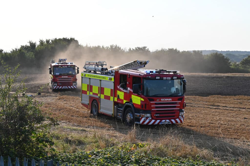 Two red Northamptonshire Fire and Rescue Service fire engines drive across a dry harvested field, raising dust behind them. The lead vehicle has blue lights illuminated, with a second fire engine following in the background. Trees line the edge of the field under a bright, clear sky.